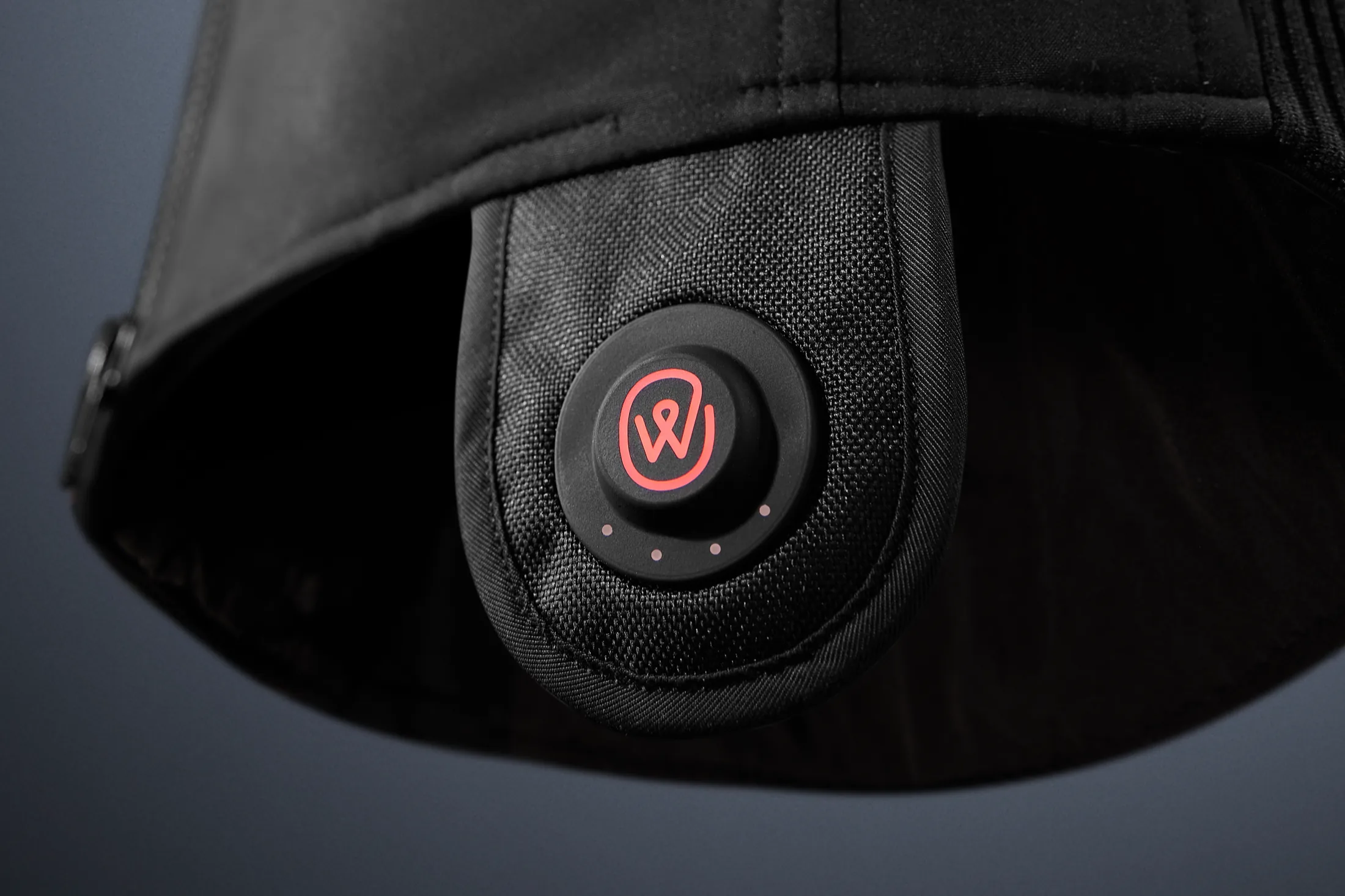 Close-up of the coral ewool mark on the round snapConnect control button, sewn into a black heated jacket above its four heat-level indicators.