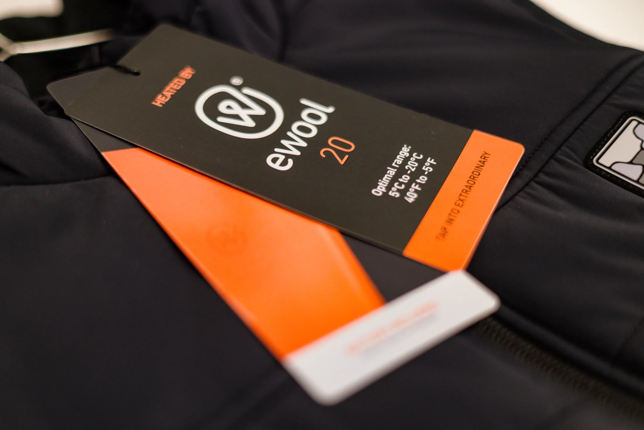 A black ewool hangtag lying on a black garment, reading Heated by ewool 20 above an optimal range of 5°C to -20°C, with an orange band down its edge set in Tap into extraordinary.