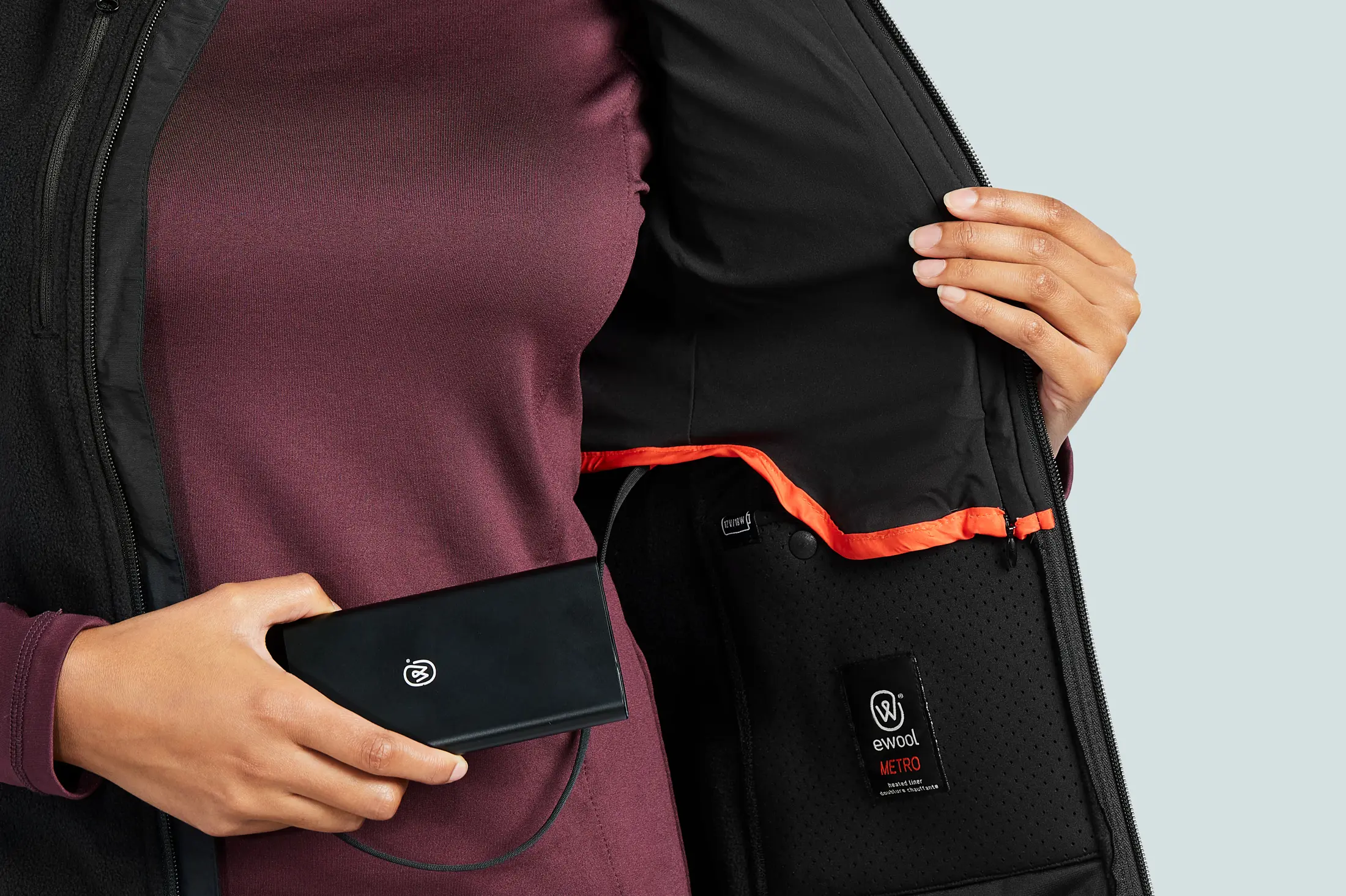 The inside of an ewool Metro heated liner held open, its perforated black lining carrying the ewool METRO heated liner label beside an orange-trimmed battery pocket, with a hand holding the connected battery pack.