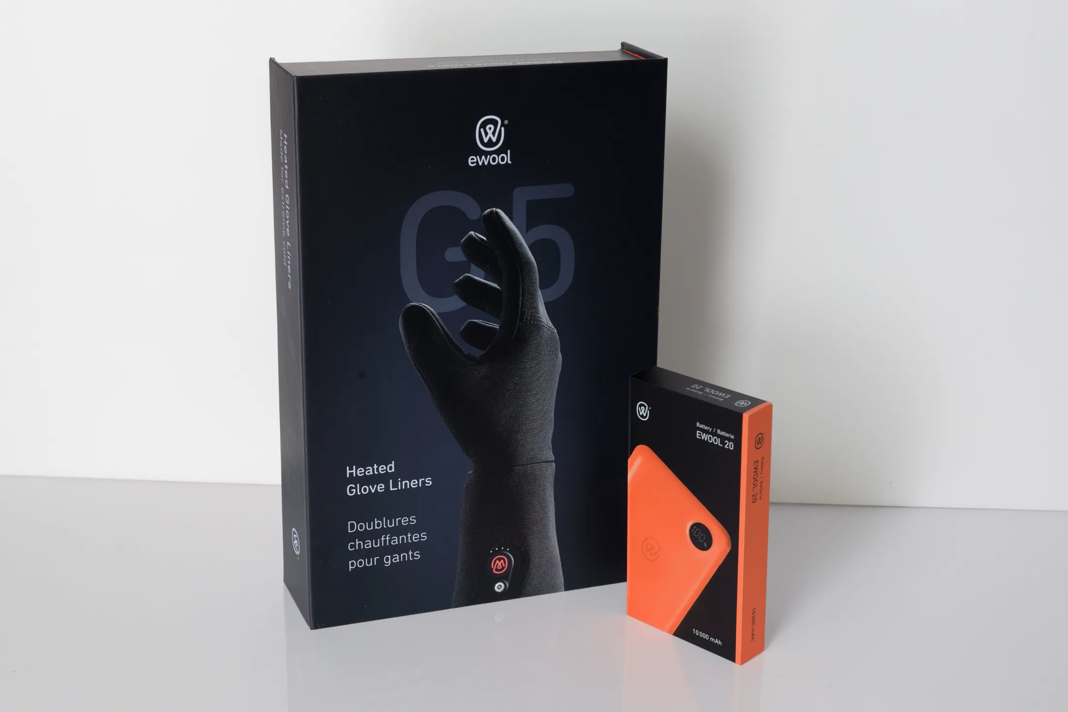 Two ewool boxes on a white surface: the tall navy G5 box for the Heated Glove Liners, printed with a black-gloved hand, and beside it the small EWOOL 20 battery box carrying the orange battery across its wrap.