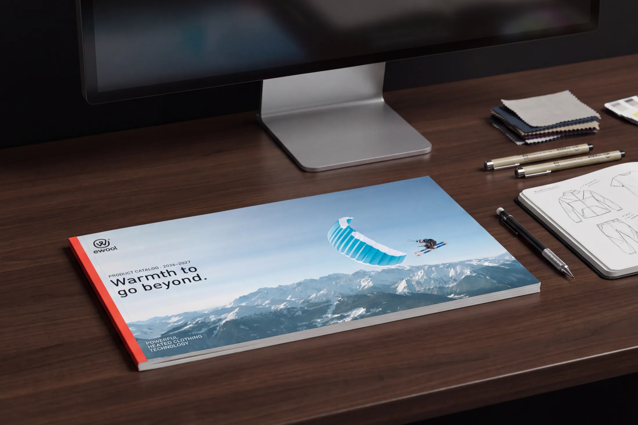 The printed ewool 2026–2027 product catalogue on a walnut desk, its cover reading Warmth to go beyond over a skier under a blue canopy above snowy peaks, beside fabric swatches and a sketchbook.