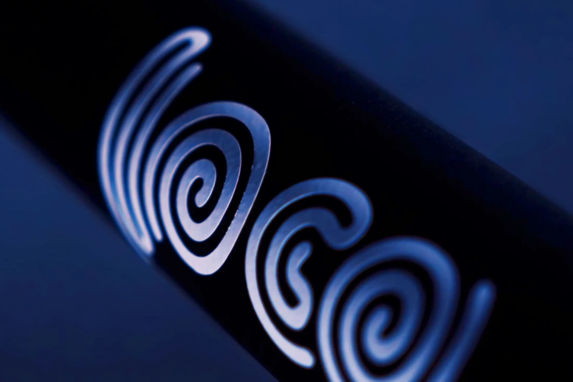 Macro of the loco wordmark, three white spirals, printed along the black stem of an electric scooter and lit deep blue from one side.