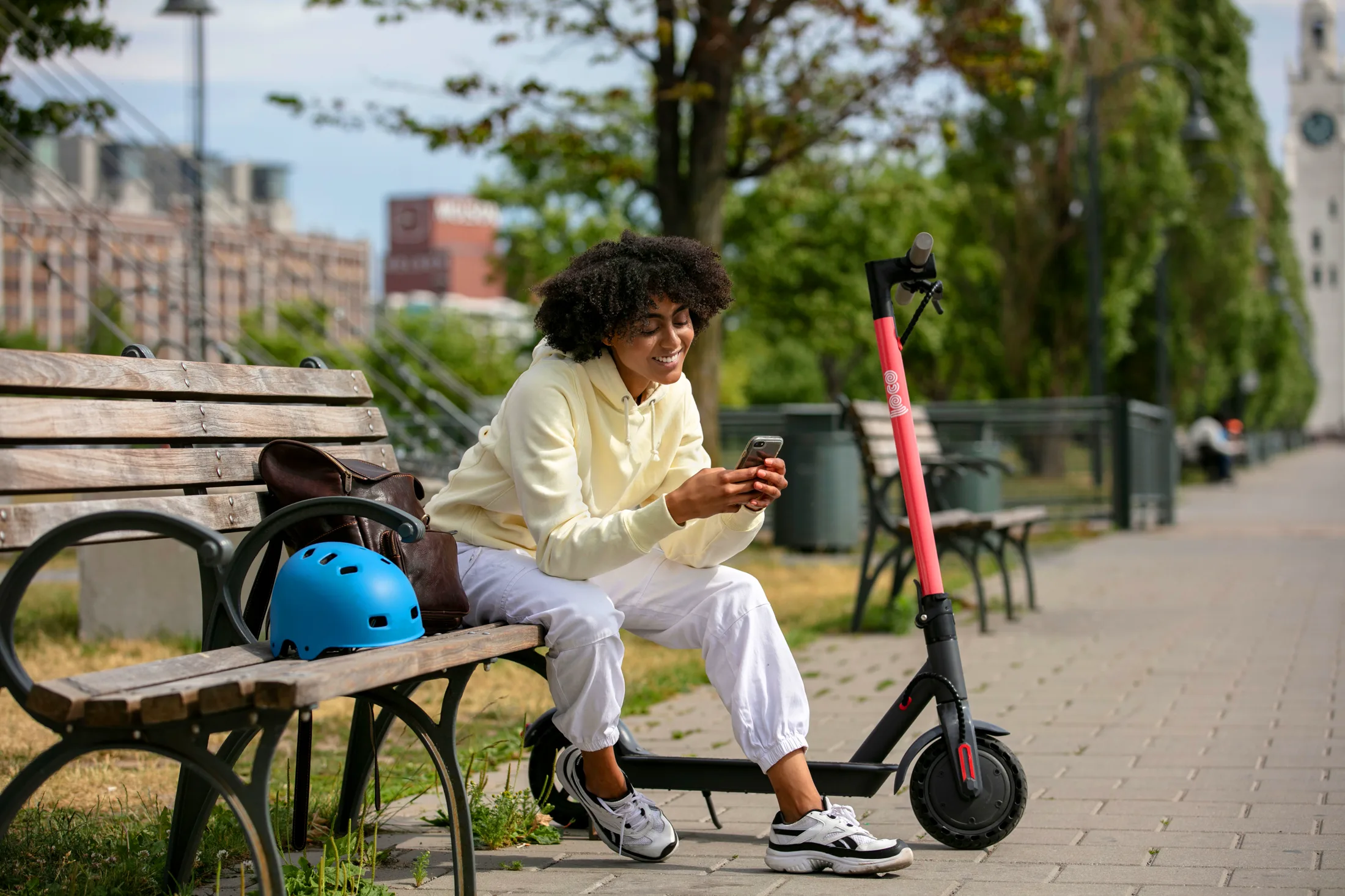 A rider sitting on a park bench looking at her phone, her Loco S1 scooter parked beside her and a blue helmet on the seat, the Old Port clock tower down the path behind.