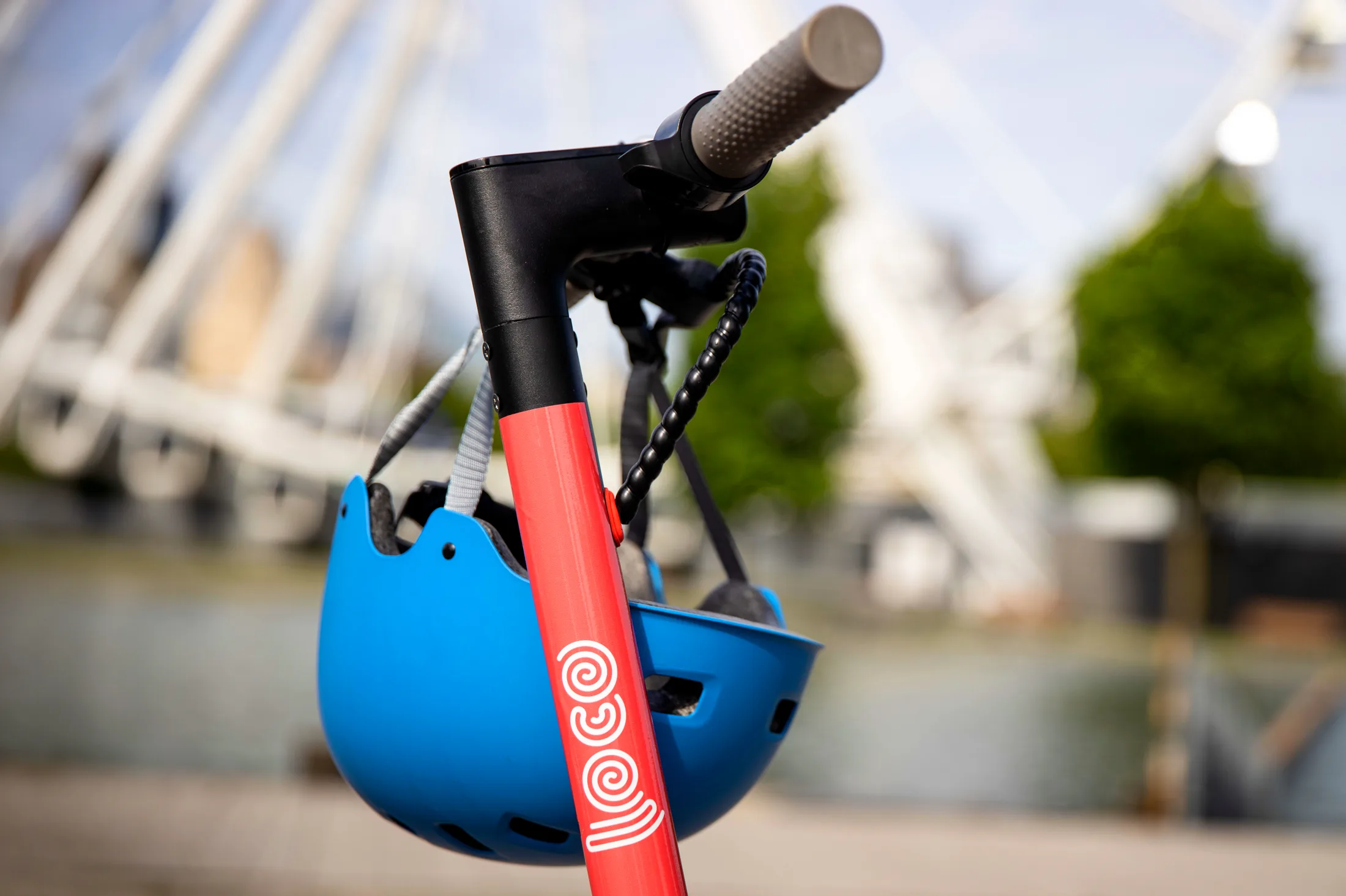 The coral stem of a Loco S1 scooter carrying the white loco mark, a blue helmet hooked over its handlebar, the Grande Roue out of focus behind it.