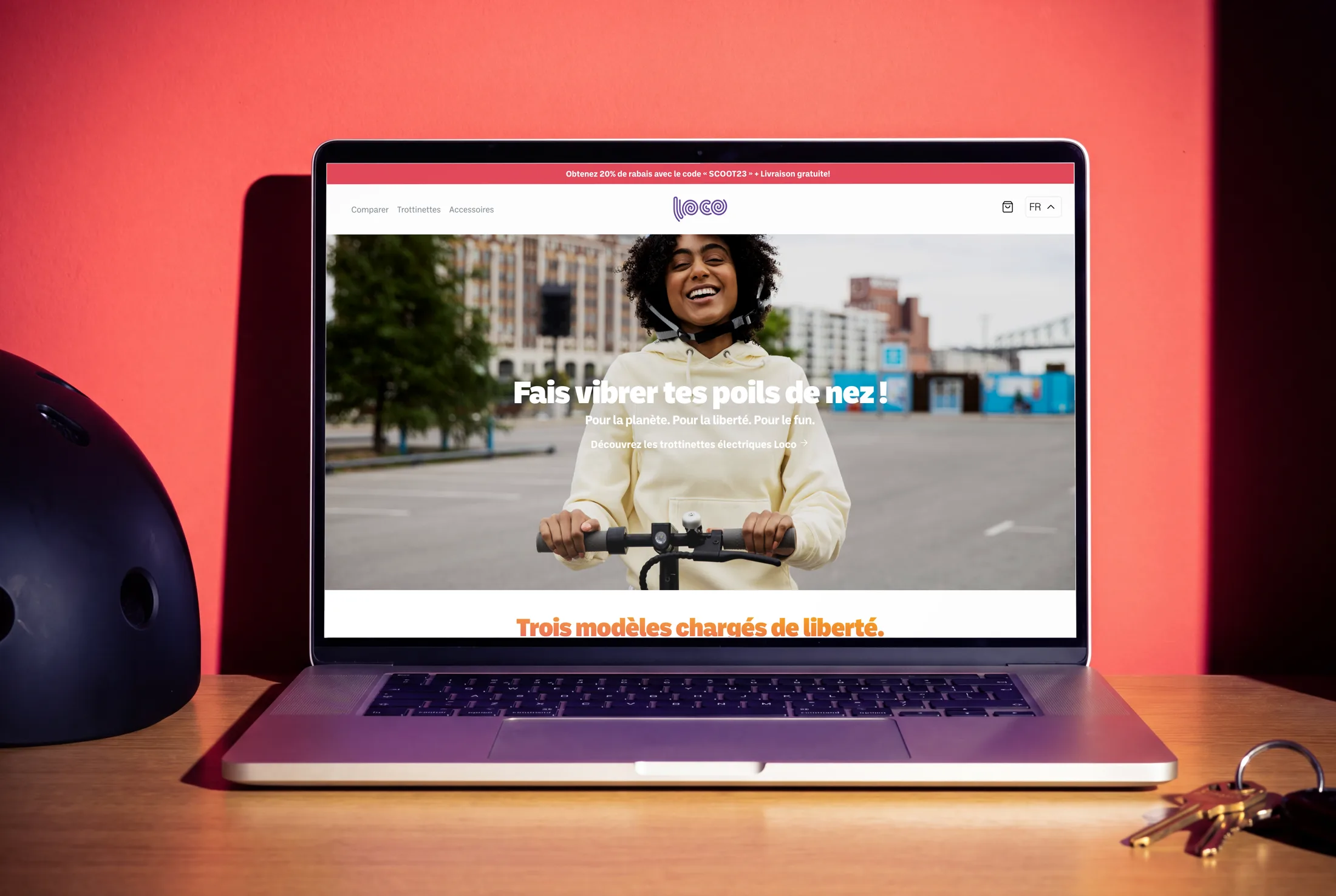 The French Loco homepage open on a laptop against a coral wall, a grinning rider behind the headline Fais vibrer tes poils de nez, with a helmet and keys on the desk.