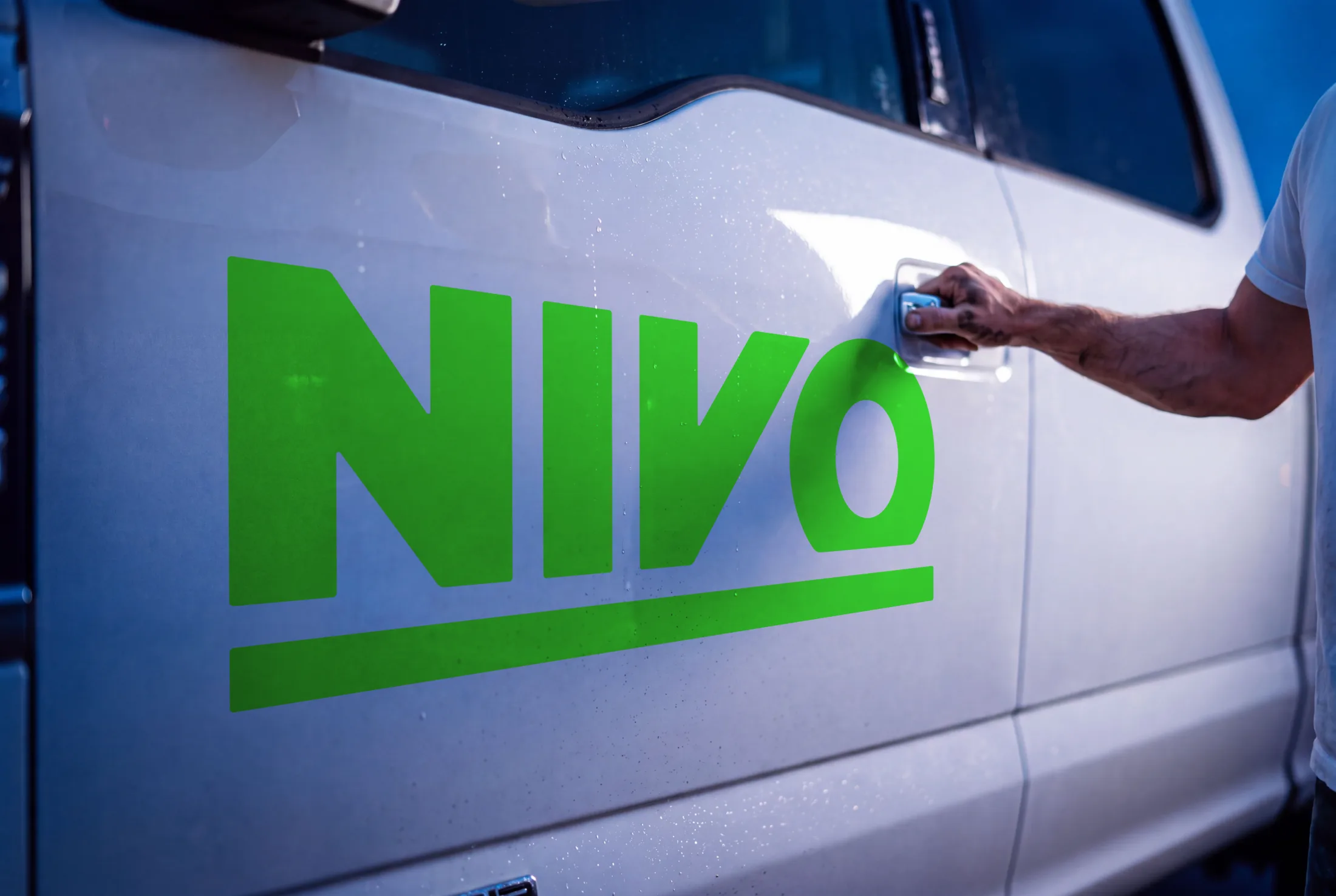 The green NIVO wordmark and its underscore rule applied to the white door of a work truck at blue hour, a hand reaching for the chrome handle beside it.
