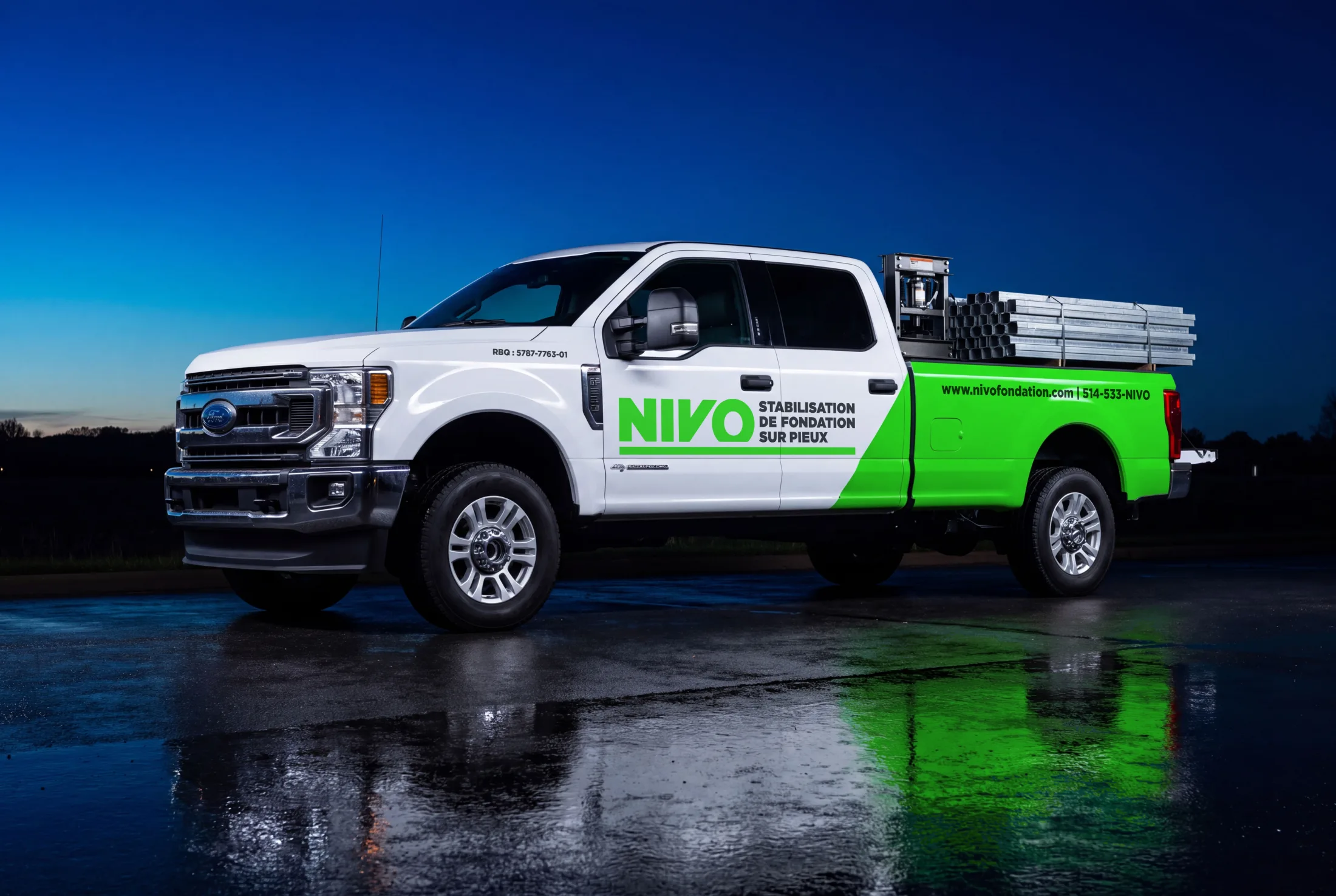 A wrapped white Ford F-250 in three-quarter view on wet asphalt under a deep blue sky, the green wedge running back over the box and galvanized pile sections loaded in the bed.