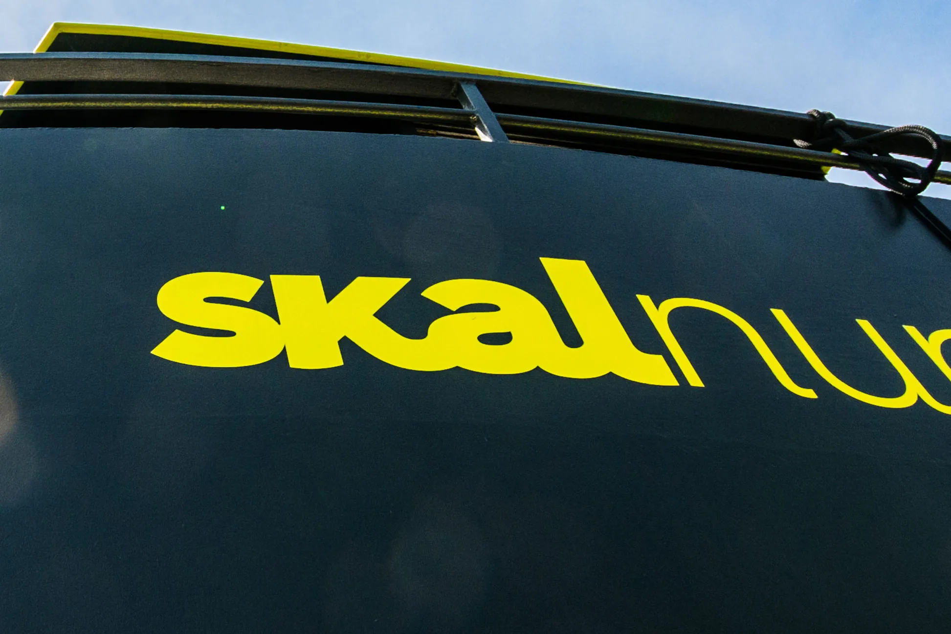 Close-up of the yellow-green skal wordmark applied to the vessel’s black hull, nuphar continuing in lighter type off the edge of the frame under the upper deck.