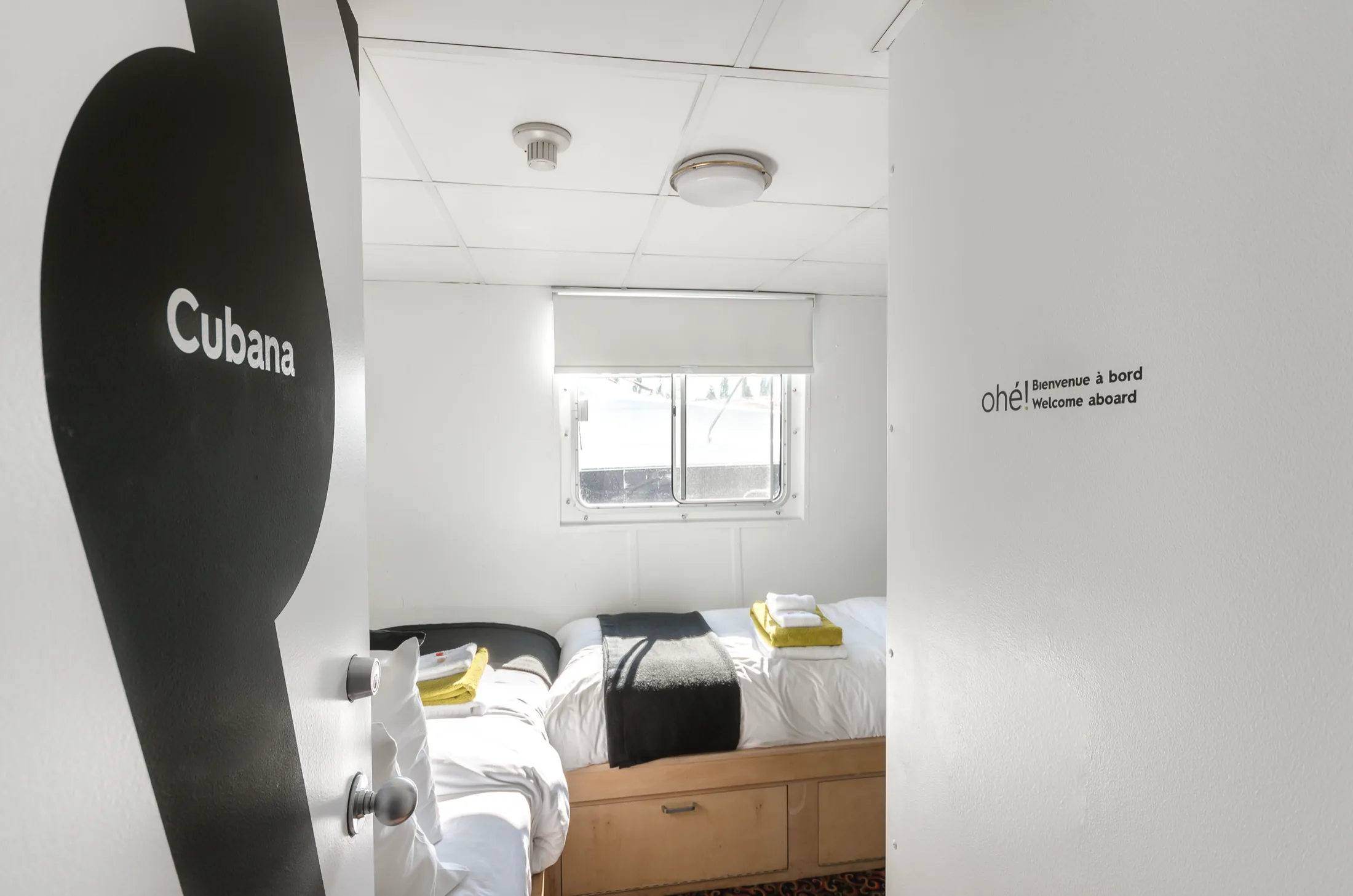 The Cubana cabin door standing open onto a white berth with folded towels on the bed, the wall opposite lettered ohé ! Bienvenue à bord / Welcome aboard.