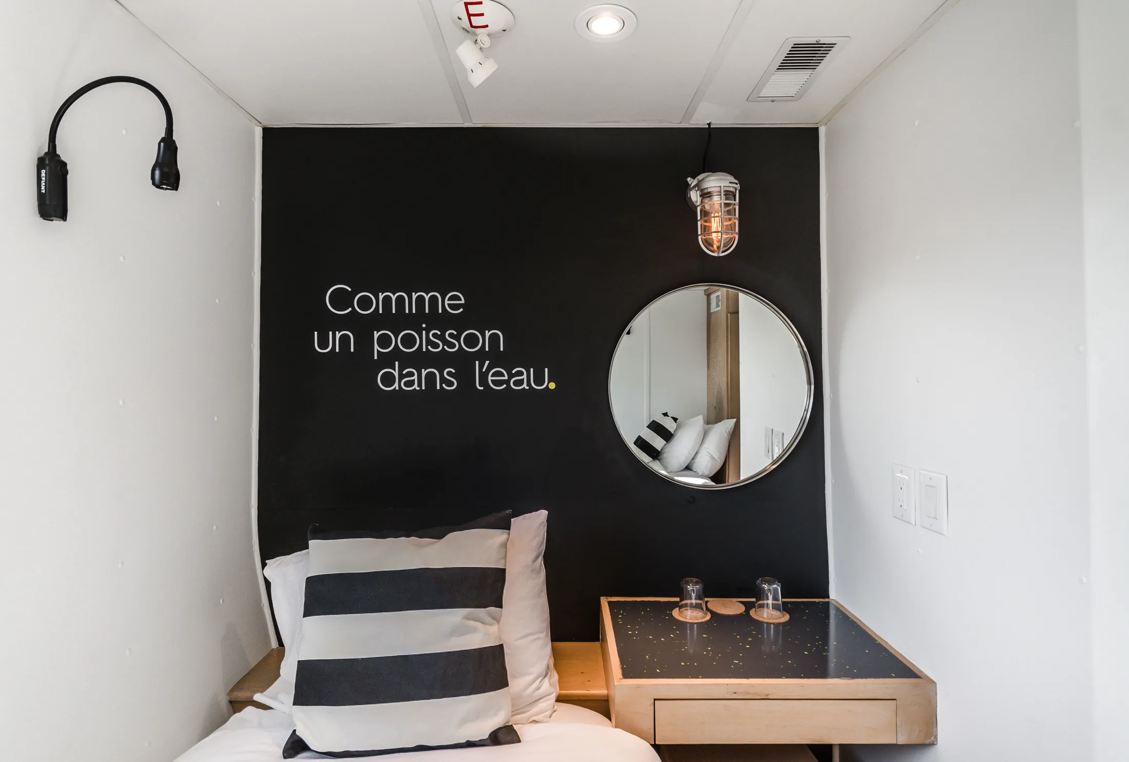 A cabin interior with a black feature wall reading Comme un poisson dans l’eau, a round mirror and a caged bulkhead light above the desk, and a striped cushion on the berth.