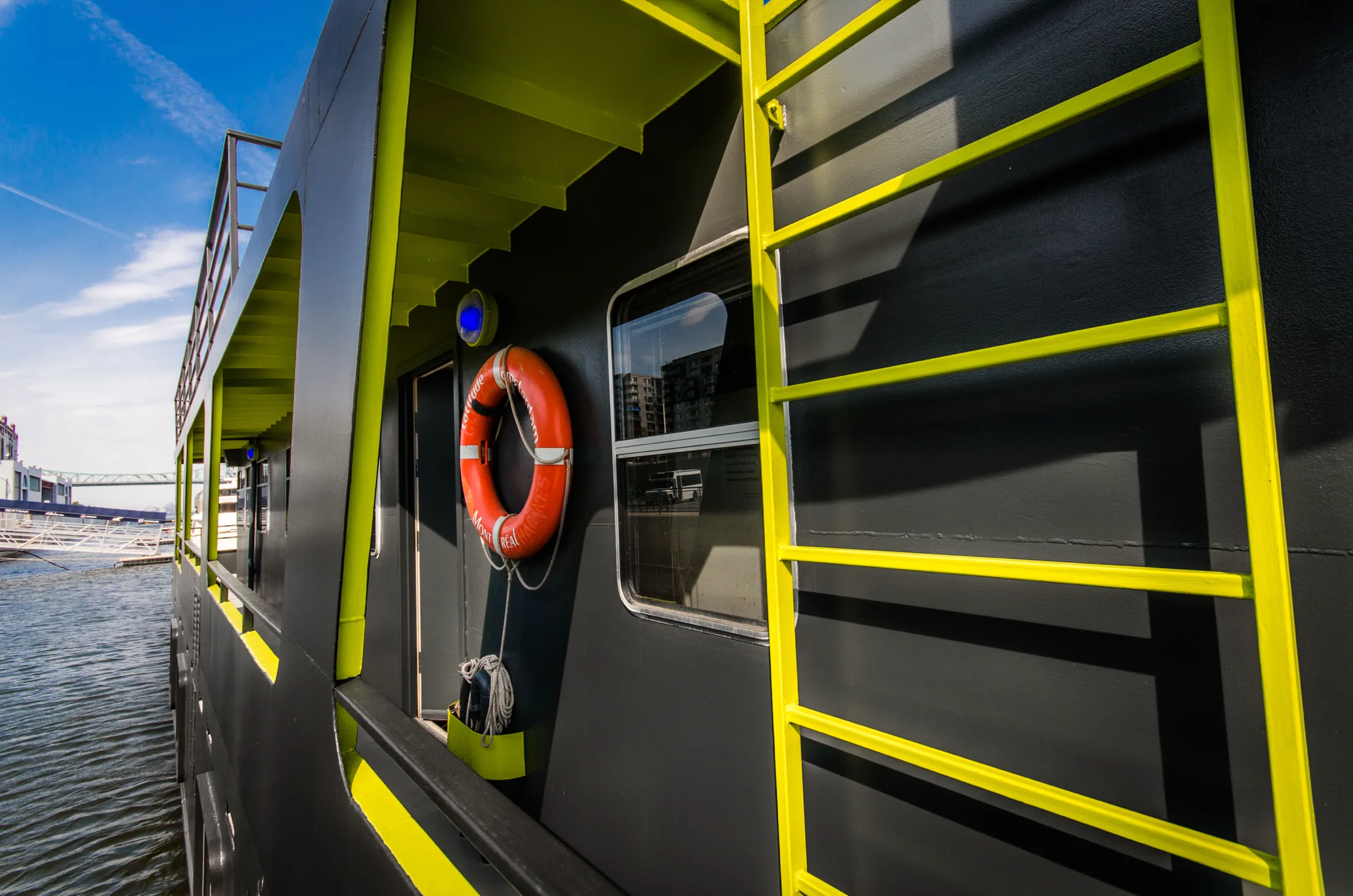 The vessel’s flank at the waterline, a yellow-green ladder painted up the black hull beside the cabin windows and an orange life ring hung outside a door.