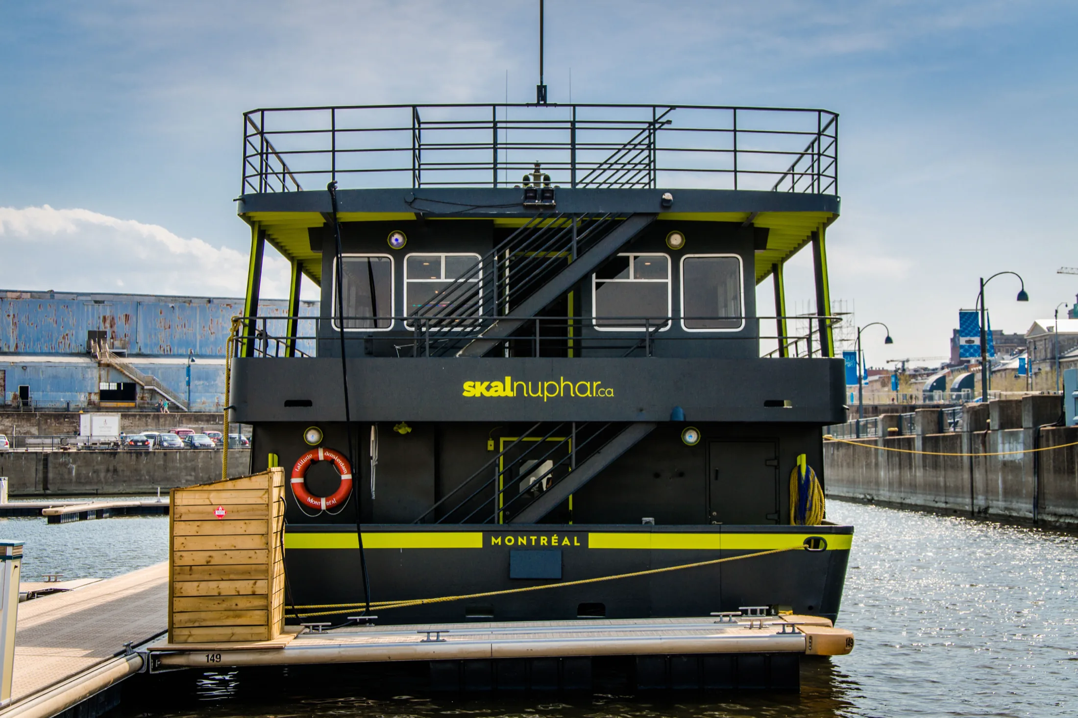 The stern of the Skal Nuphar from the dock, skalnuphar.ca set in yellow-green across the transom above a band reading MONTRÉAL, with the gangway and an orange life ring at the boarding deck.