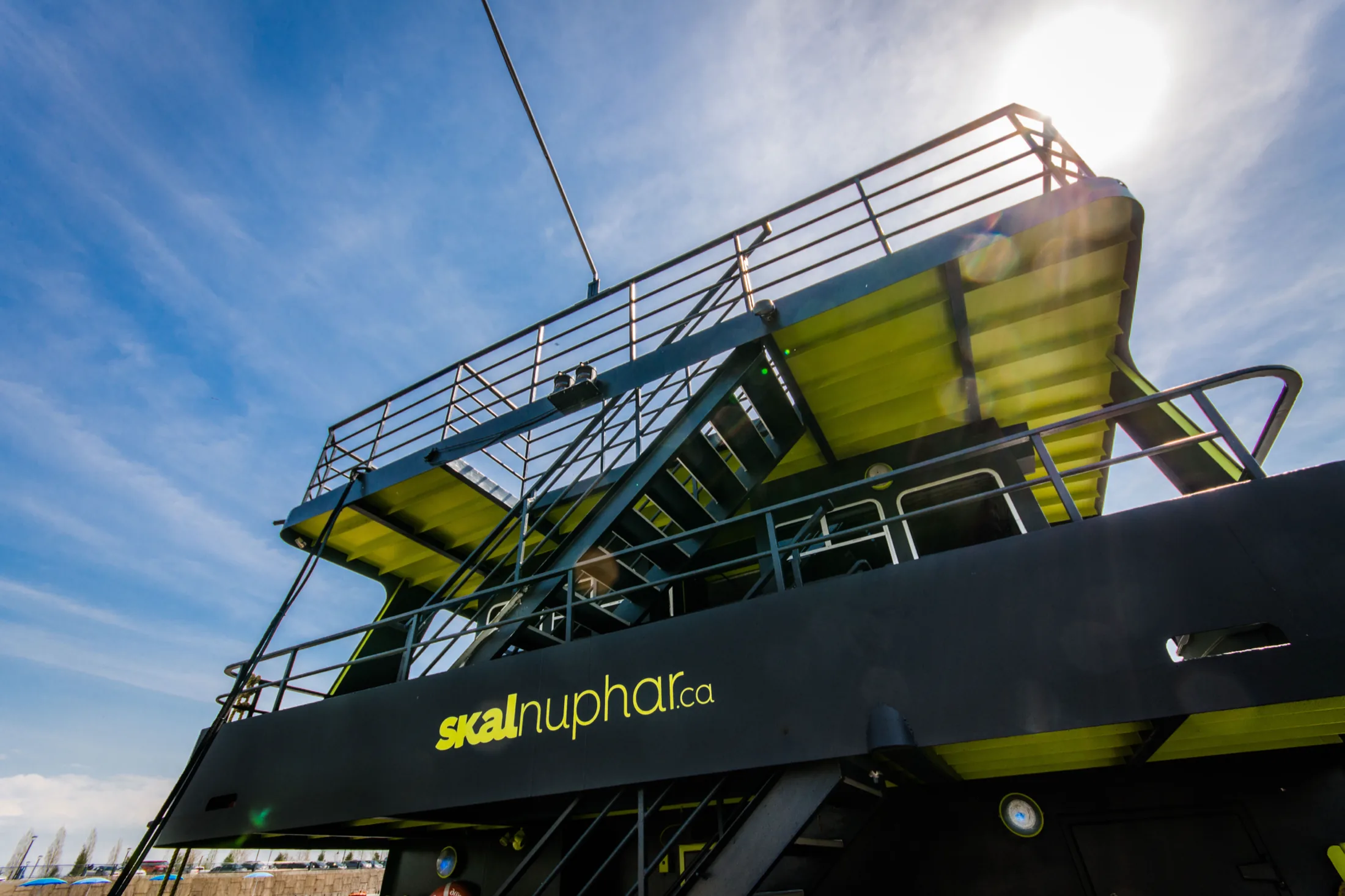 Low-angle view of the vessel’s upper decks against the sun, the brand’s yellow-green carried across the underside of every ceiling and the skalnuphar.ca mark on the black hull below.